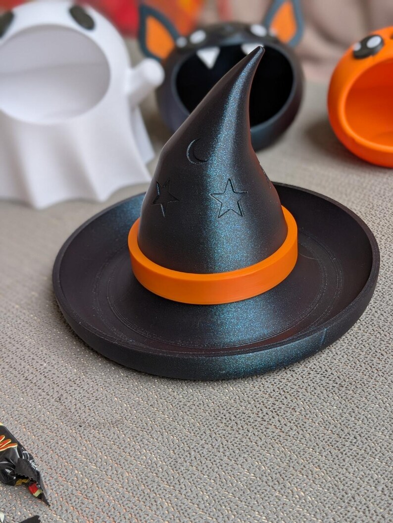 May include: A black witch hat-shaped trinket dish with an orange band, moon, and star cutouts. Other Halloween-themed containers are in the background, including a ghost, bat, and pumpkin.