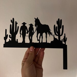 May include: Black metal silhouette of a couple walking a horse in a desert landscape with cacti. The silhouette is mounted on a metal bracket.