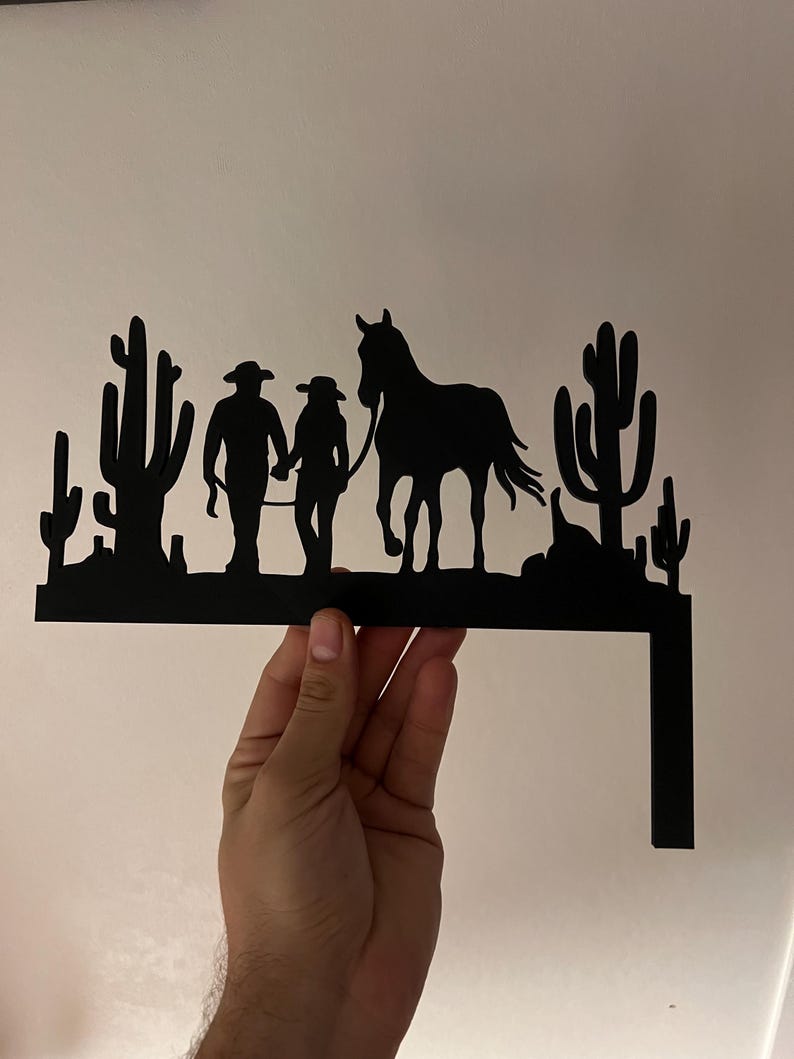 May include: Black metal silhouette of a couple walking a horse in a desert landscape with cacti. The silhouette is mounted on a metal bracket.