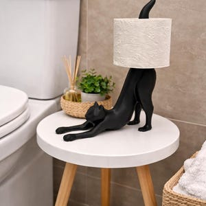 Pode incluir: Um porta-papel higi&ecirc;nico em forma de gato preto. O gato est&aacute; em posi&ccedil;&atilde;o de alongamento, sustentando um rolo de papel higi&ecirc;nico branco estampado. O suporte est&aacute; sobre uma mesa lateral branca com pernas de madeira, para decora&ccedil;&atilde;o de banheiro.