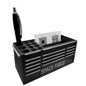 May include: Black desk organizer with silver accents, featuring the text "SPACE FORCE". It includes a pen, multiple small circular holes, and a slot for business cards. The design resembles a miniature toolbox.