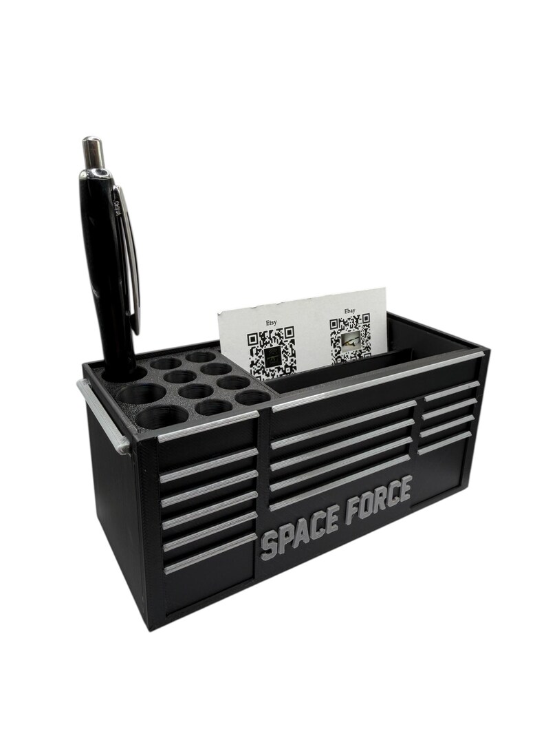 May include: Black desk organizer with silver accents, featuring the text "SPACE FORCE". It includes a pen, multiple small circular holes, and a slot for business cards. The design resembles a miniature toolbox.