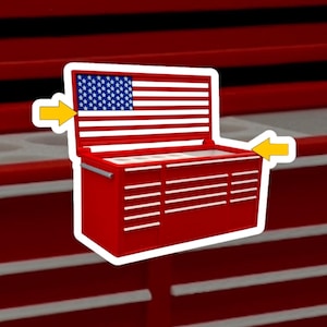 May include: A red metal tool chest sticker featuring a white border and two yellow arrows pointing toward the unit. The open lid displays a graphic of the American flag with white stars on a blue field and horizontal white stripes on a red background.
