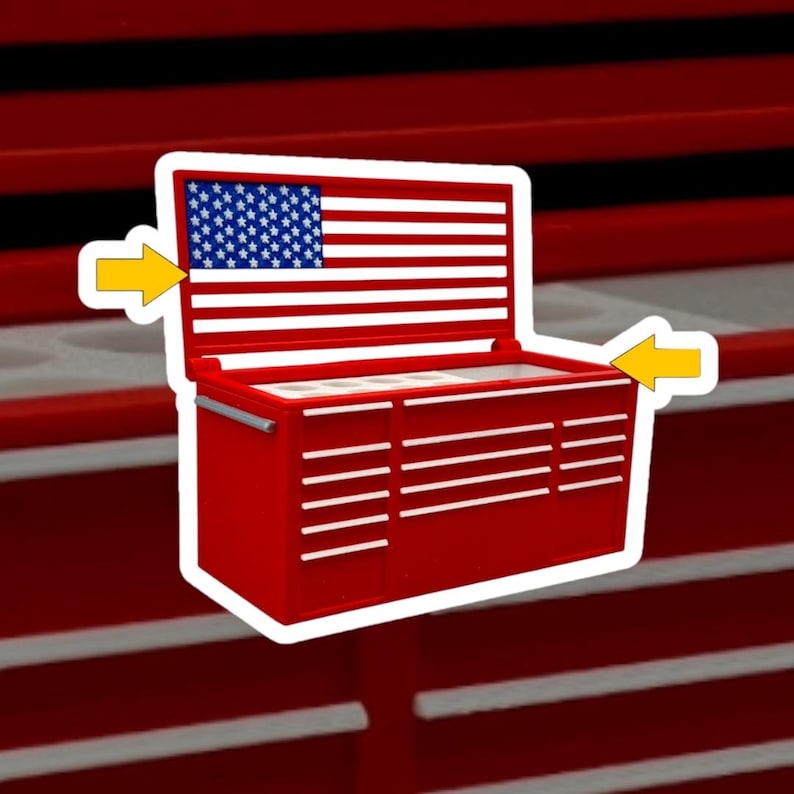 May include: A red metal tool chest sticker featuring a white border and two yellow arrows pointing toward the unit. The open lid displays a graphic of the American flag with white stars on a blue field and horizontal white stripes on a red background.