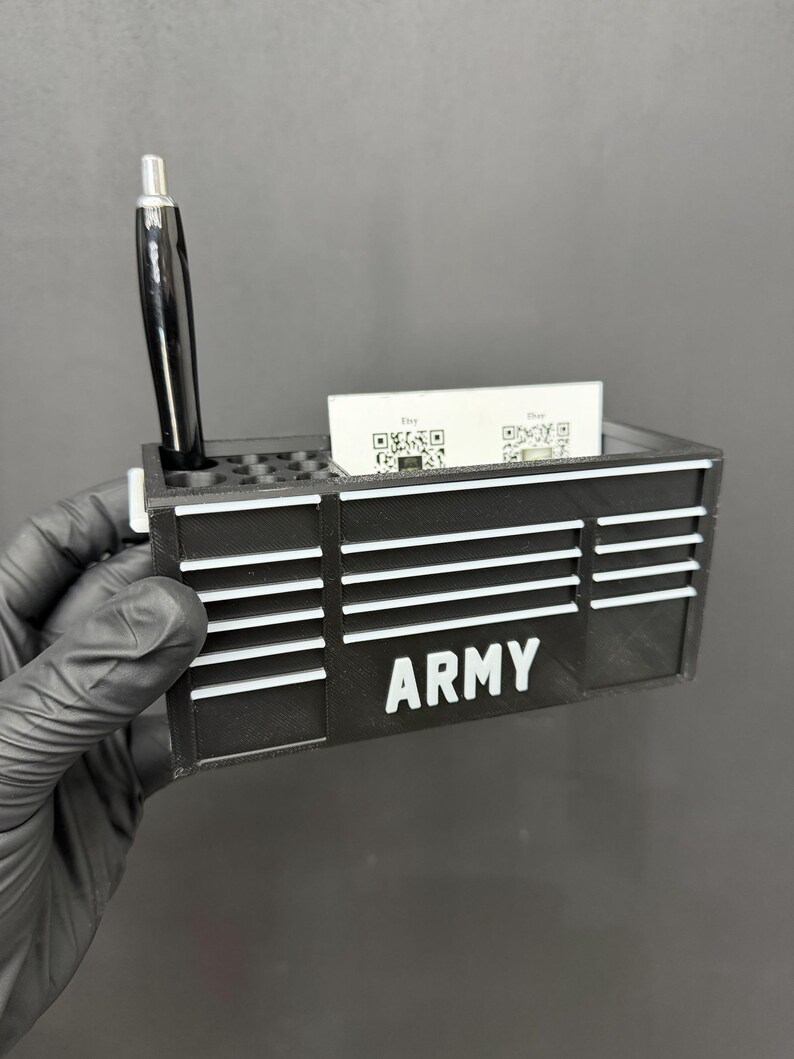 May include: Black desk organizer with the word "ARMY" in white block letters. The organizer has slots for pens and business cards. A black pen is in the pen slot. Business cards with QR codes are in the card slot.