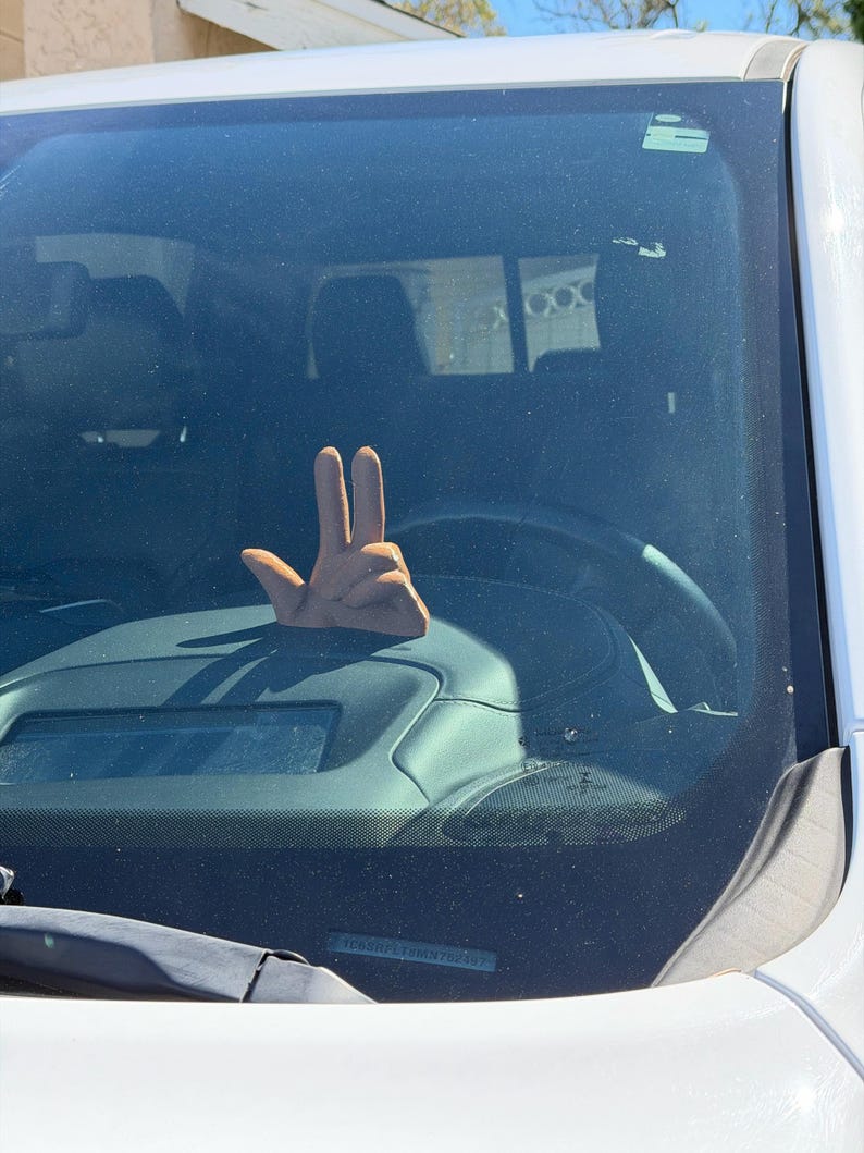 Puede incluir: Un accesorio para coche en forma de mano marr&oacute;n, con los dedos &iacute;ndice y coraz&oacute;n extendidos, se ve a trav&eacute;s de un parabrisas. El accesorio est&aacute; en el salpicadero, cerca del volante. El coche es blanco.