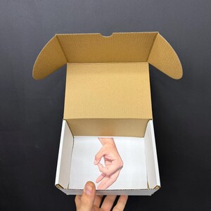 May include: An open cardboard box with a white interior and a brown exterior. The inside of the box features a printed image of a hand making the 'okay' gesture. The box is held by a hand against a black background.
