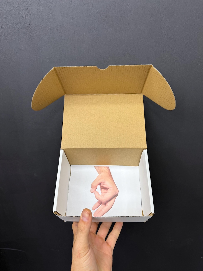 May include: An open cardboard box with a white interior and a brown exterior. The inside of the box features a printed image of a hand making the 'okay' gesture. The box is held by a hand against a black background.