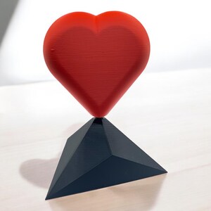 Pu&ograve; includere: Una scultura a forma di cuore rosso si trova su una base triangolare nera. Il cuore &egrave; liscio e lucido, mentre la base &egrave; opaca.