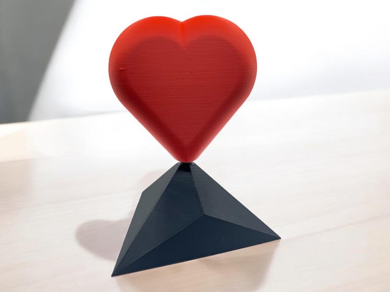 Pu&ograve; includere: Una scultura a forma di cuore rosso si trova su una base triangolare nera. Il cuore &egrave; liscio e lucido, mentre la base &egrave; opaca.