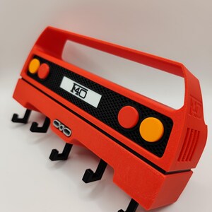 May include: A red wall-mounted key holder shaped like a car's rear end, featuring black hooks for keys. The design includes orange and red circular lights, a black mesh area, and the number "F40" in white. The key holder is made of plastic.