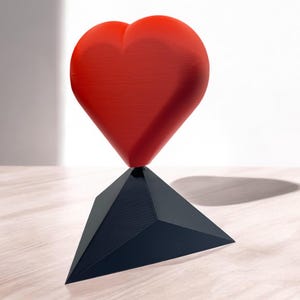 Pu&ograve; includere: Una scultura a forma di cuore rosso si trova su una base triangolare nera. La scultura &egrave; realizzata in un materiale liscio e lucido.