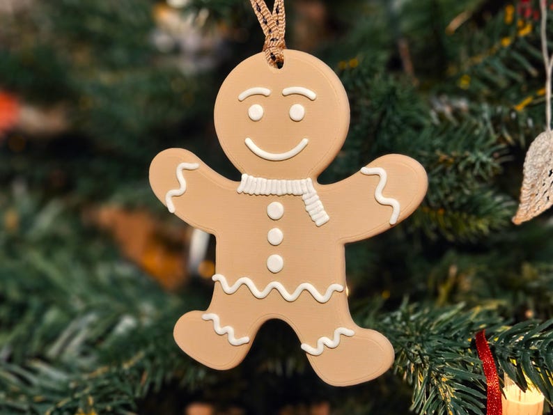 May include: A tan gingerbread man Christmas ornament with white icing details, including a scarf, buttons, and wavy lines. The ornament is hanging in front of a blurred green Christmas tree.