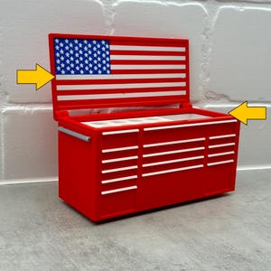 May include: A red miniature tool chest with a hinged lid featuring a blue, white, and red American flag design. The chest has multiple white drawer handles and a silver side handle. Two yellow arrows point to the lid and the open top compartment.