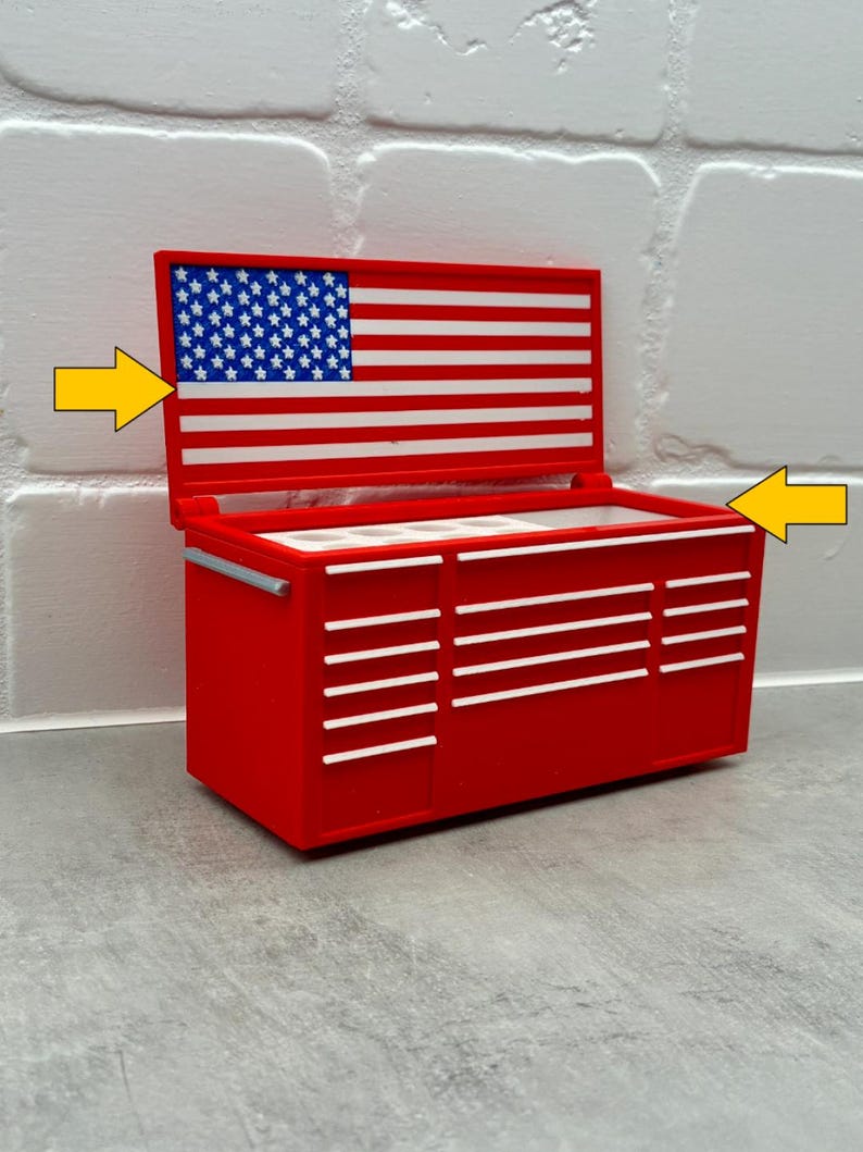 May include: A red miniature tool chest with a hinged lid featuring a blue, white, and red American flag design. The chest has multiple white drawer handles and a silver side handle. Two yellow arrows point to the lid and the open top compartment.