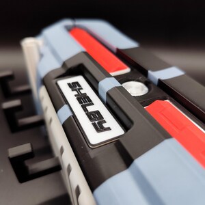 K&ouml;nnte beinhalten: Nahaufnahme eines mehrfarbigen Objekts mit dem Namen "SHELBY" in wei&szlig;en Buchstaben. Das Objekt weist blaue, schwarze, rote und wei&szlig;e Elemente auf. Es scheint ein Bestandteil eines gr&ouml;&szlig;eren Ger&auml;ts oder Modells zu sein.