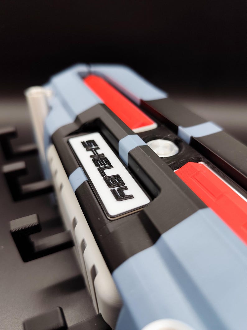 K&ouml;nnte beinhalten: Nahaufnahme eines mehrfarbigen Objekts mit dem Namen "SHELBY" in wei&szlig;en Buchstaben. Das Objekt weist blaue, schwarze, rote und wei&szlig;e Elemente auf. Es scheint ein Bestandteil eines gr&ouml;&szlig;eren Ger&auml;ts oder Modells zu sein.