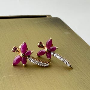 May include: A pair of gold-toned earrings featuring a floral design with ruby-colored petals and a stem of small, clear gemstones. The earrings are set on a gold surface.