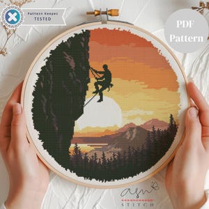 Op de afbeelding: Een borduurring met kruissteek met een bergbeklimmersscène bij zonsondergang. Het ontwerp toont een silhouet van een klimmer, een grote zon en een landschap van bergen en bomen. De tekst "PDF Pattern" is zichtbaar.