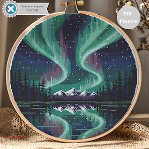 Peut inclure: Une broderie au point de croix des aurores boréales, avec des teintes vertes et violettes sur un ciel bleu foncé. Le motif comprend une forêt, des montagnes et un lac réfléchissant. La broderie est placée dans un cercle en bois. Le texte "Pattern Keeper TESTED" est visible.