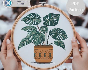 May include: A cross-stitch pattern for a potted monstera plant. The plant is in a brown pot with a geometric pattern. The pattern is in a hoop and being held by a person's hands. The text "PDF Pattern" is in the upper right corner of the image.