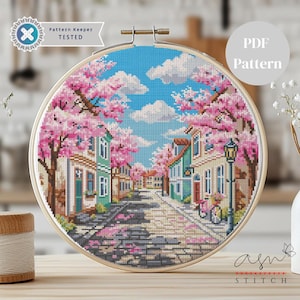 May include: Cross-stitch pattern of a street scene with buildings and cherry blossom trees in full bloom. The design is framed in a wooden embroidery hoop. The sky is blue with fluffy white clouds. Text on the image reads "Pattern Keeper TESTED" and "PDF Pattern".