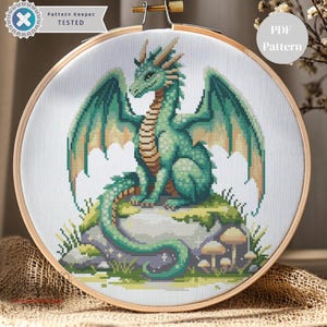 Puede incluir: Un patrón de punto de cruz de un dragón verde con alas y escamas detalladas, posado sobre una roca cubierta de musgo. El diseño está enmarcado en un bastidor de bordado de madera. El texto "Pattern Keeper TESTED" y "PDF Pattern" son visibles.