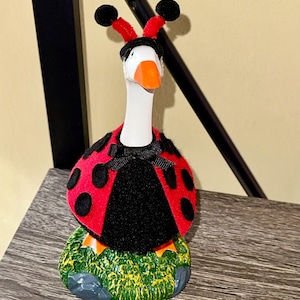 May include: A whimsical goose figurine dressed as a ladybug. The goose has a white neck and orange beak, wearing a red and black spotted costume with a black bow. It stands on a green and gray base, with red antennae and black pom-poms.