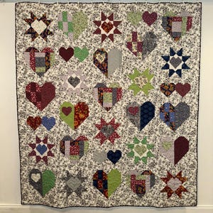 May include: A quilt with a white background and a variety of colorful patchwork hearts and stars. The hearts are made of different fabrics and patterns, and the stars are all red with white points.
