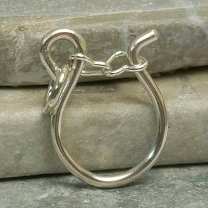 Puede incluir: Un anillo plateado con un diseño único y en bucle. El anillo presenta un gran lazo en forma de lágrima conectado a un lazo más pequeño y circular y un elemento curvo en forma de gancho. El metal tiene una superficie pulida y reflectante.