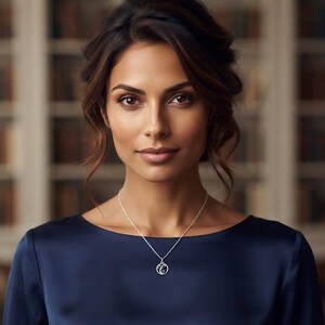 May include: A silver necklace featuring a circular pendant with an intricate script letter C, suspended on a delicate chain. The pendant is polished and reflective, resting against a dark navy blue satin top with a wide, rounded neckline.