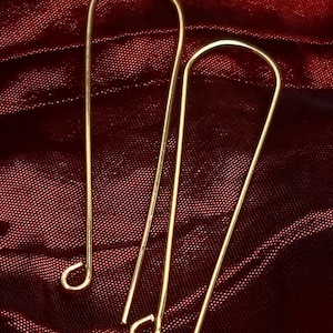 May include: A pair of gold-colored, elongated earrings. Each earring has a curved top and a small loop at the bottom. The earrings are displayed against a dark red, textured background.