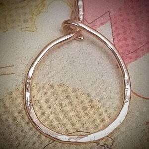 May include: A hammered metal pendant in a teardrop shape. The pendant is a light, warm color, possibly rose gold or copper. The design features a loop at the top for a chain. The metal has a textured, uneven surface.