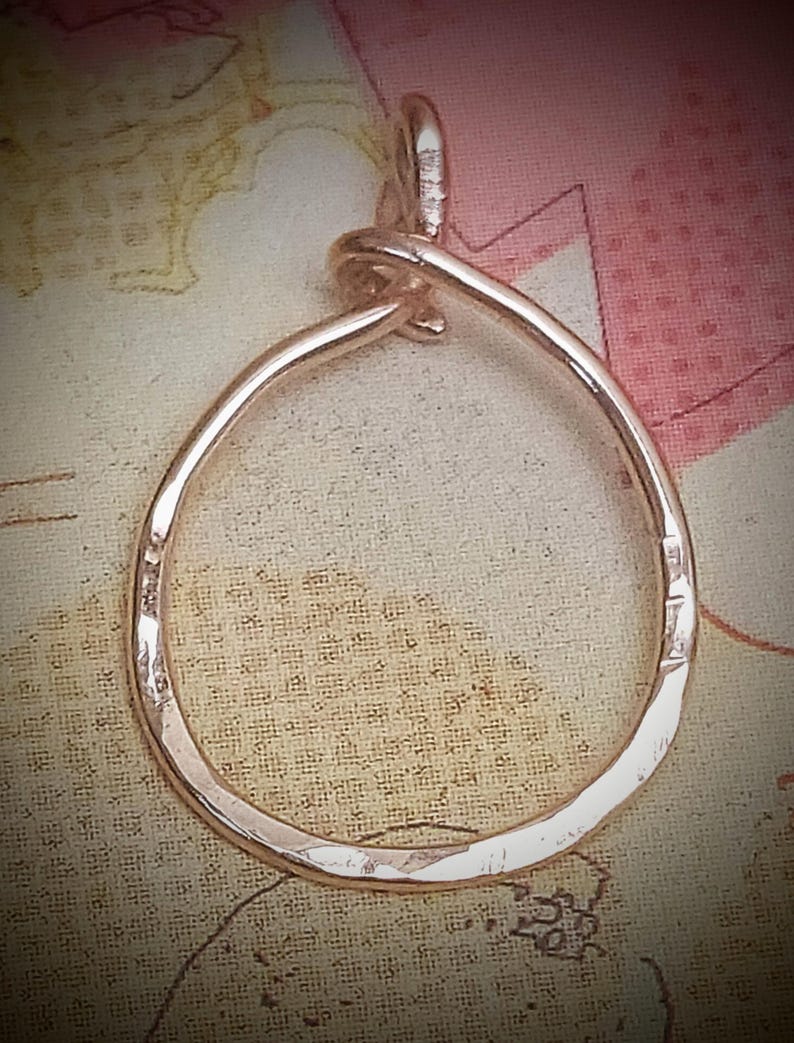 May include: A hammered metal pendant in a teardrop shape. The pendant is a light, warm color, possibly rose gold or copper. The design features a loop at the top for a chain. The metal has a textured, uneven surface.