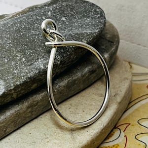 May include: A silver-colored, open-circle bracelet with a knot-like design at the top. The bracelet is made of a shiny, reflective metal and is displayed on a gray stone surface. The design is simple and elegant.