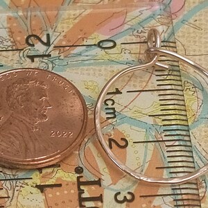 May include: A copper penny next to a small, hammered metal hoop pendant. The penny, dated 2022, is next to the pendant, which is approximately 1 inch in diameter. The pendant is on a patterned surface with a ruler.