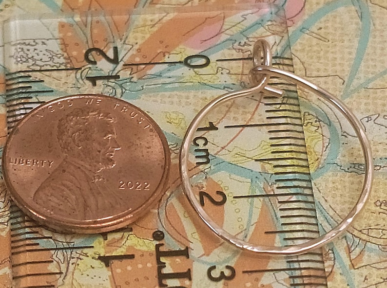 May include: A copper penny next to a small, hammered metal hoop pendant. The penny, dated 2022, is next to the pendant, which is approximately 1 inch in diameter. The pendant is on a patterned surface with a ruler.