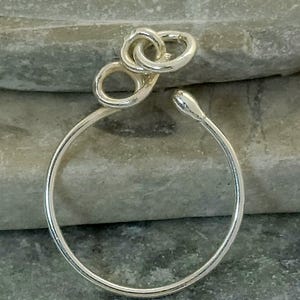 May include: A silver-colored hoop earring with a unique design. The earring features a circular hoop with an open end and a decorative knot-like element at the top. The earring is shown against a gray stone background.