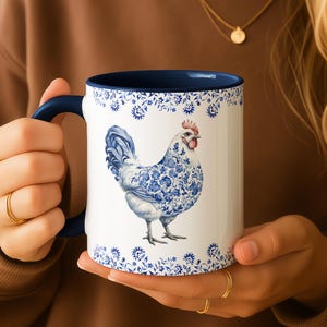 May include: White ceramic mug with a blue interior and handle. The mug features a detailed illustration of a hen with blue floral patterns. The design includes a blue floral border. A gold necklace is visible in the background.