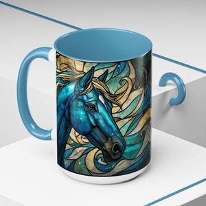 May include: A white ceramic mug with a blue handle and interior. The mug features a detailed illustration of a blue horse head with a golden mane and a stained-glass effect. The background has swirling gold and teal accents.