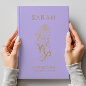 May include: A light purple hardcover journal featuring gold foil text and graphics. The cover displays the name SARAH at the top, a floral illustration intertwined with the Capricorn zodiac symbol in the center, and the words CAPRICORN Love, Devotion, Loyalty.