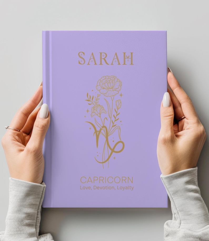 May include: A light purple hardcover journal featuring gold foil text and graphics. The cover displays the name SARAH at the top, a floral illustration intertwined with the Capricorn zodiac symbol in the center, and the words CAPRICORN Love, Devotion, Loyalty.