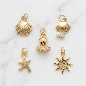 May include: A collection of five gold-toned metal charms featuring nautical and celestial designs. The set includes a ridged seashell, a small octopus, a seashell with a white pearl and clear stone, a textured starfish, and a spiral sun shape.