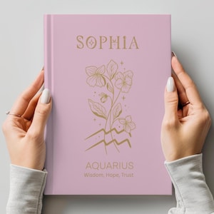 May include: A light pink hardcover journal featuring the name SOPHIA in gold lettering at the top. Below the name is a gold line art illustration of flowers and the Aquarius zodiac symbol. The text AQUARIUS and Wisdom, Hope, Trust appear below.