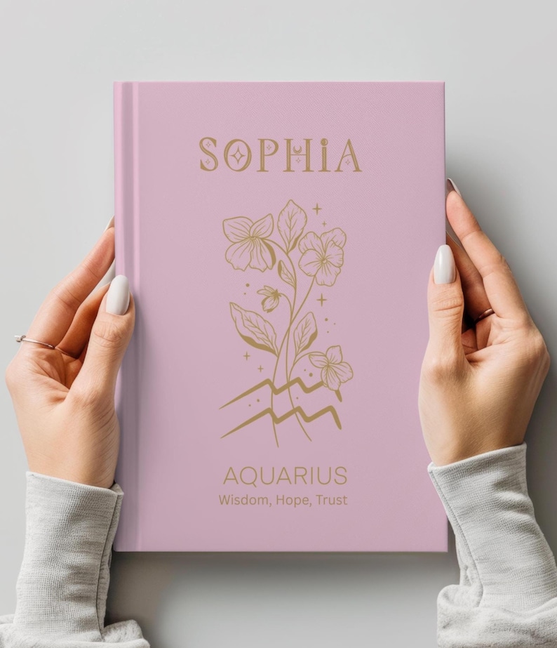 May include: A light pink hardcover journal featuring the name SOPHIA in gold lettering at the top. Below the name is a gold line art illustration of flowers and the Aquarius zodiac symbol. The text AQUARIUS and Wisdom, Hope, Trust appear below.
