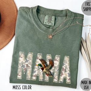May include: A moss-colored t-shirt with the word "MAMA" in a camouflage pattern, featuring a detailed illustration of a duck. A gold necklace and a brown hat are also visible. The shirt is labeled "Made in USA".
