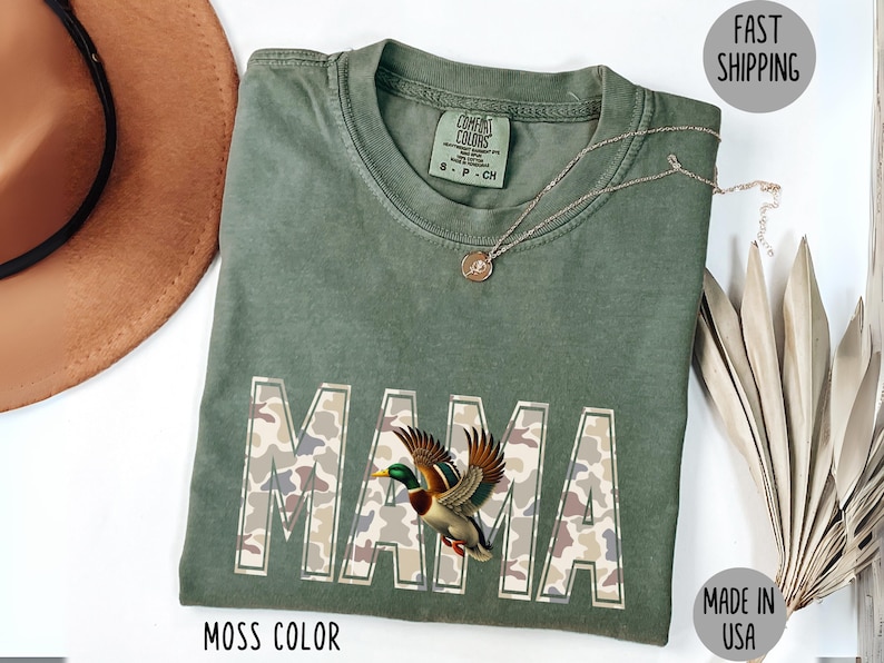 May include: A moss-colored t-shirt with the word "MAMA" in a camouflage pattern, featuring a detailed illustration of a duck. A gold necklace and a brown hat are also visible. The shirt is labeled "Made in USA".