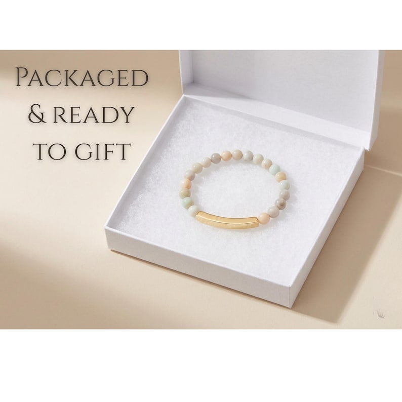 May include: A bracelet with neutral-toned beads and a gold-coloured bar is presented in an open white gift box. The text "PACKAGED & READY TO GIFT" is visible. The bracelet is designed for gifting.