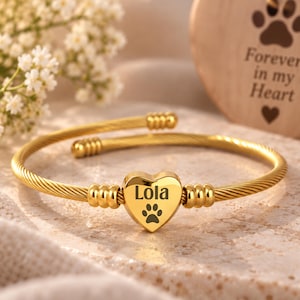May include: A gold-toned cable wire bangle bracelet featuring a central heart-shaped charm. The charm is engraved with the name Lola and a paw print. The bracelet has ribbed cylindrical accents on either side of the heart and open ends.