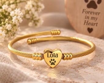 May include: A gold-toned cable wire bangle bracelet featuring a central heart-shaped charm. The charm is engraved with the name Lola and a paw print. The bracelet has ribbed cylindrical accents on either side of the heart and open ends.